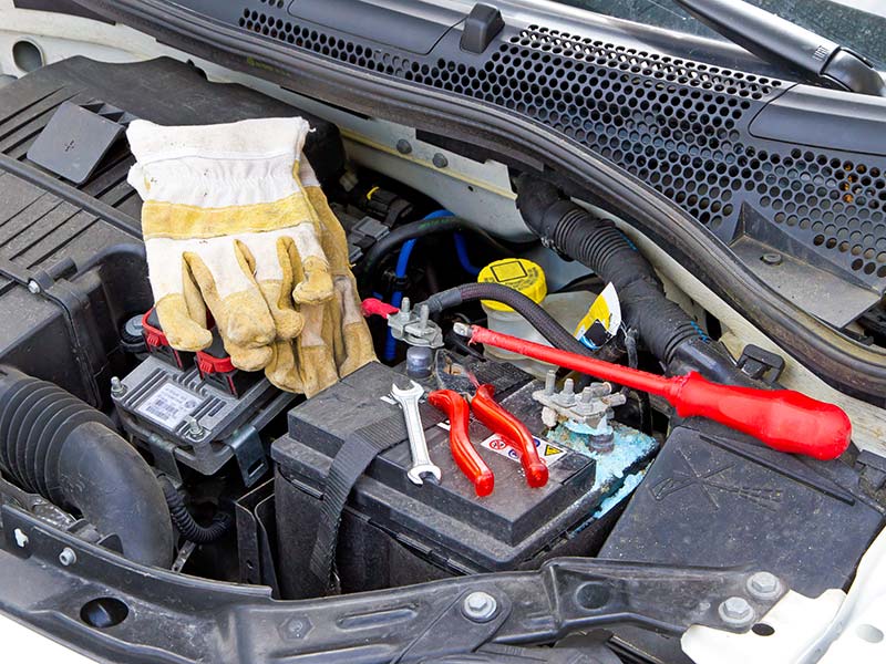 Handschuhe, Schraubendreher und anderes Werkzeug liegt neben einer Autobatterie bei geöffneter Motorhaube.