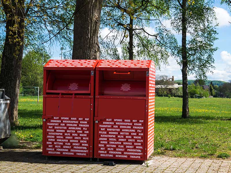 Zwei Altkleidercontainer stehen neben einem Park.