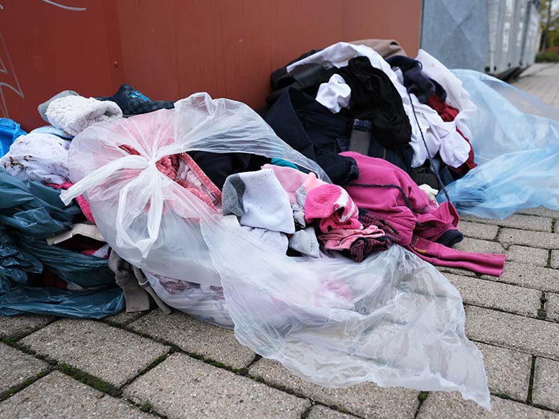 Textilien liegen zur Entsorgung in einer aufgerissenen Tüte neben einem Altkleidercontainer.